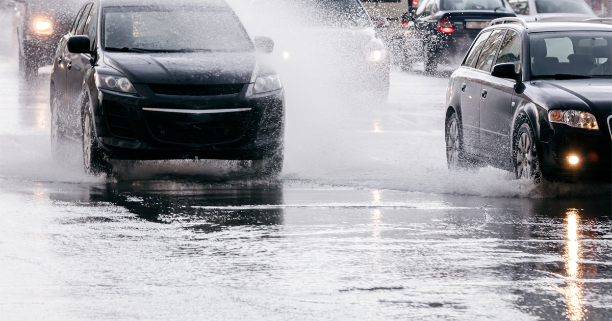 Cars driving through standing water on a flooded roadway during heavy rain, illustrating the dangers of driving on flooded roads in Michigan.